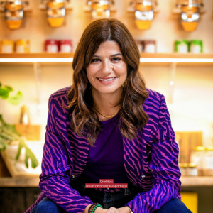 Joana Teixeira sentada num restaurante, sorridente, com blazer roxo e padrão, representando a sua marca como consultora de restaurantes sustentáveis.